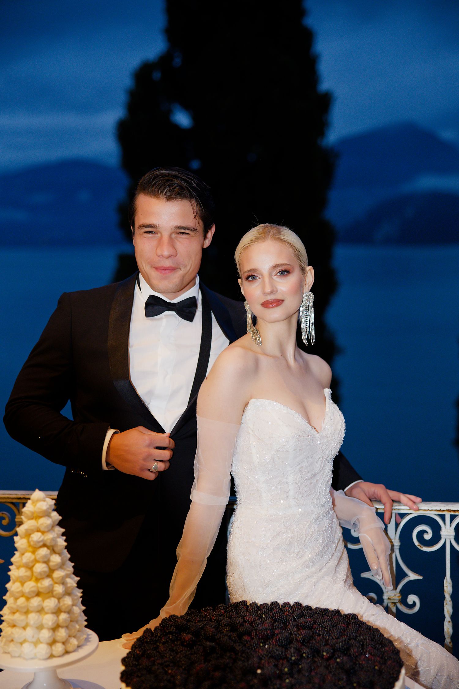 Elegant couple stands with their wedding cakes against dramatic blue twilight backdrop.