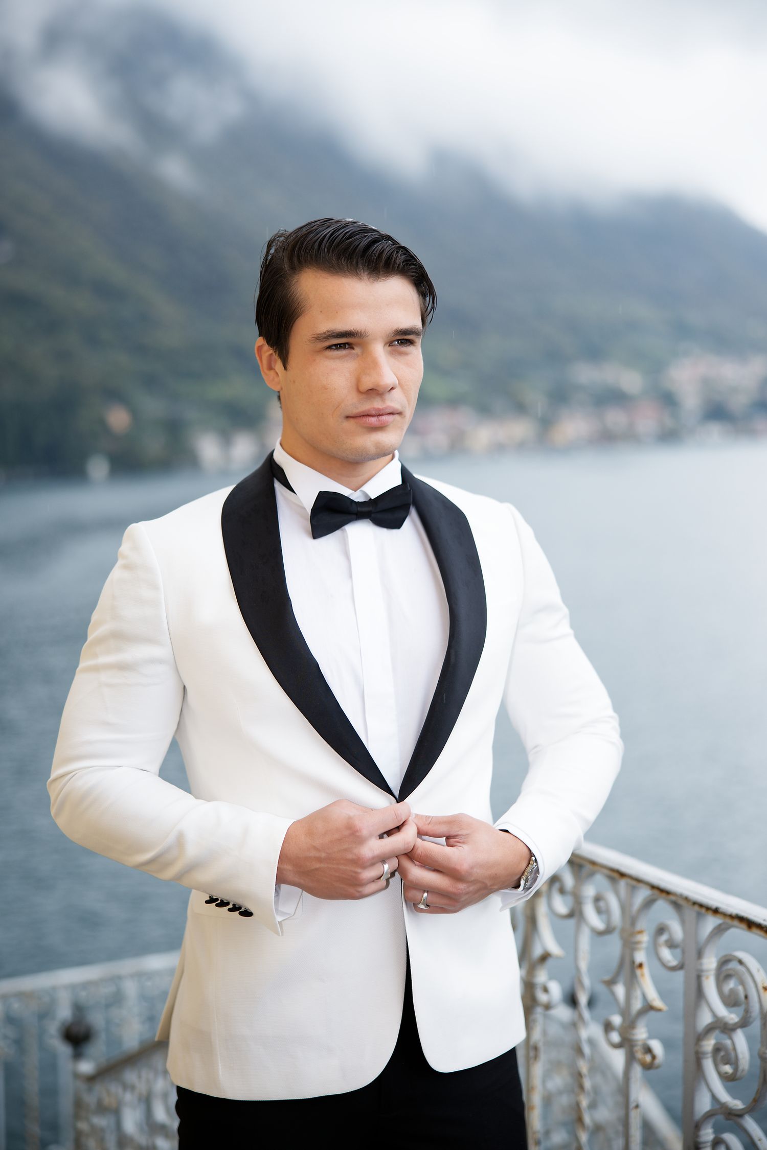 A groom in a white dinner jacket with black lapels poses against a lakeside mountain backdrop.