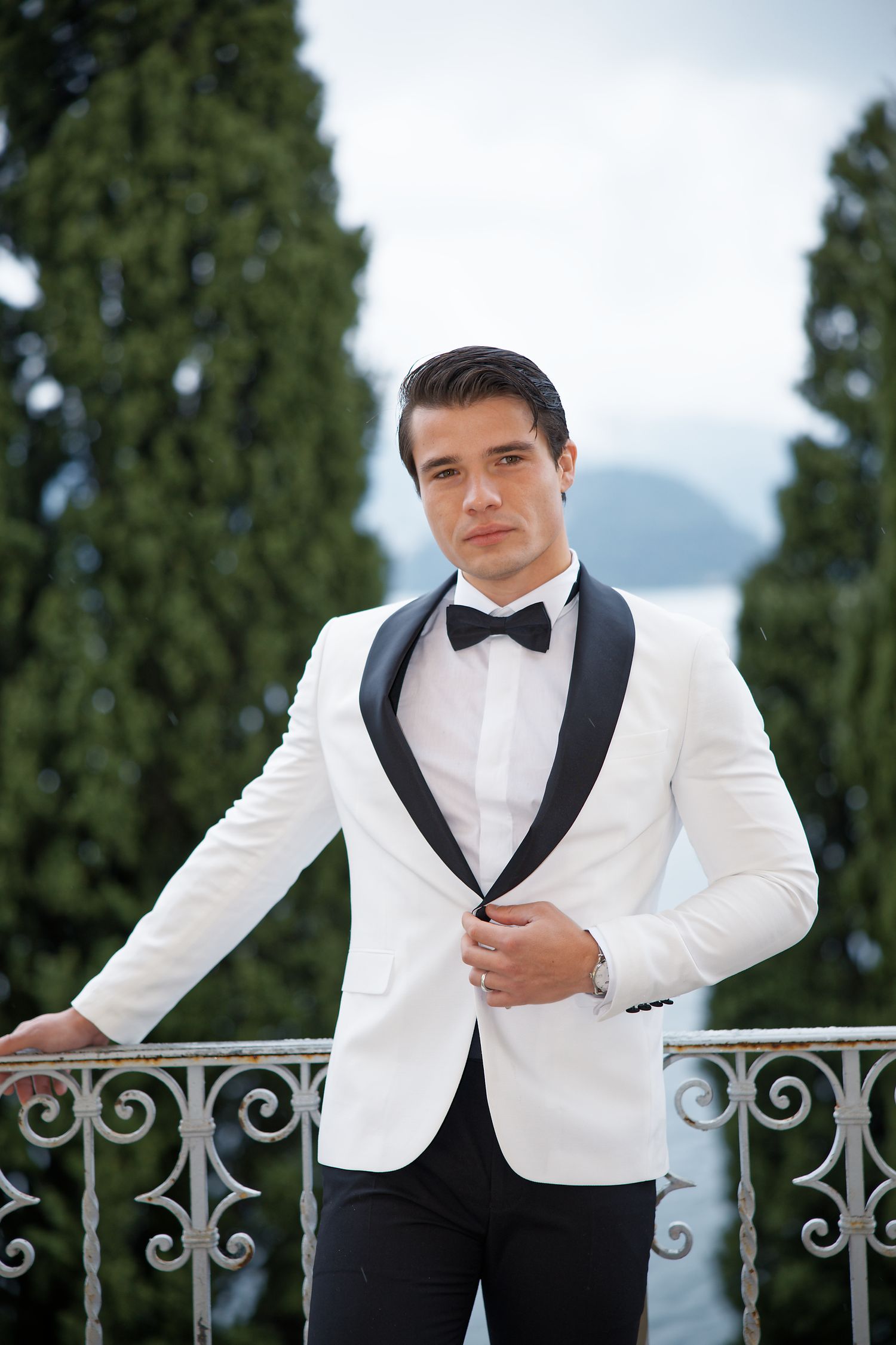 A formal portrait of a groom in white tuxedo jacket against a scenic mountain backdrop.