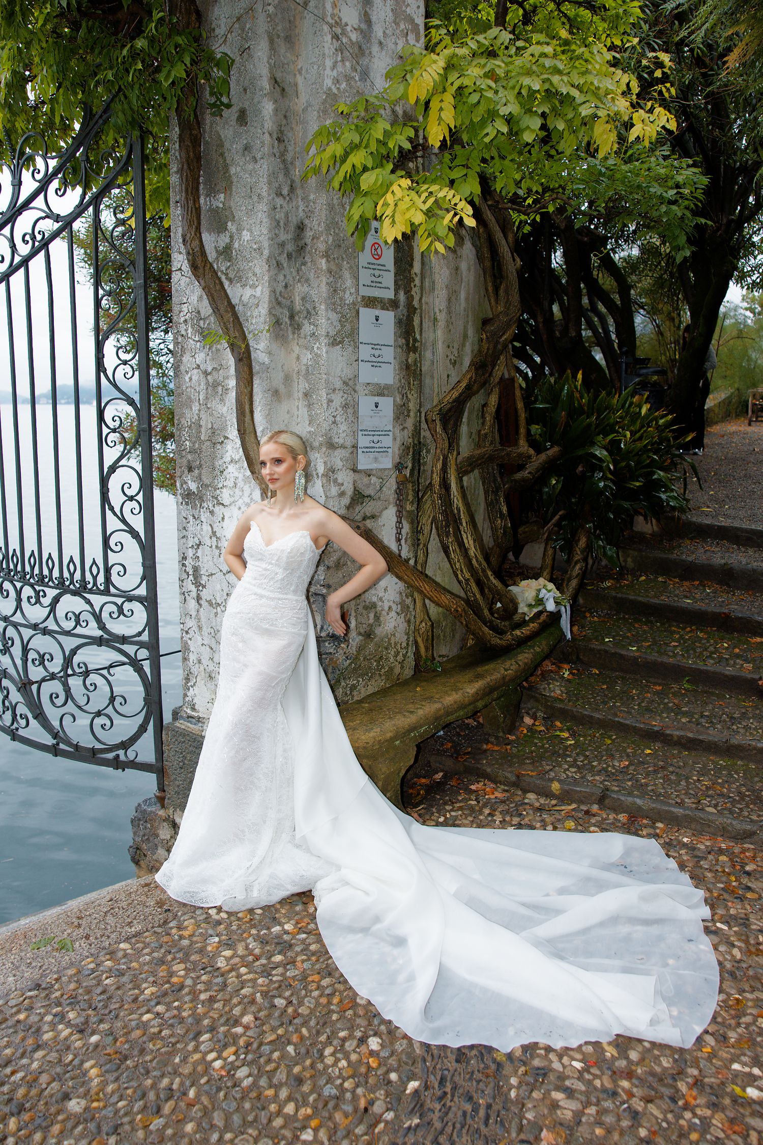 Bride in elegant train dress poses against iron gates and stone wall by lake.