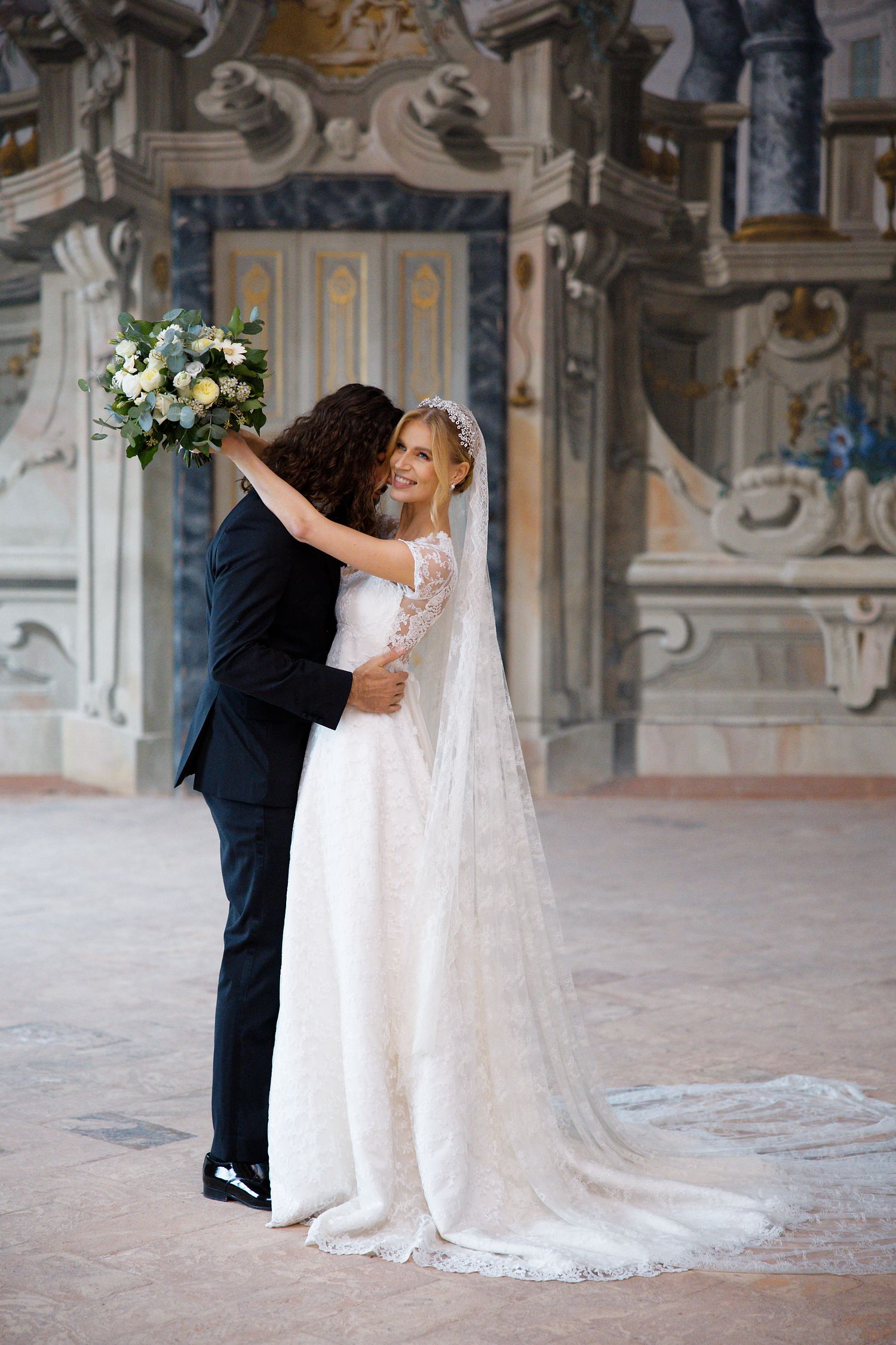 A bride in a white lace gown with long veil holds a bouquet while dancing with groom in front of ornate architecture.