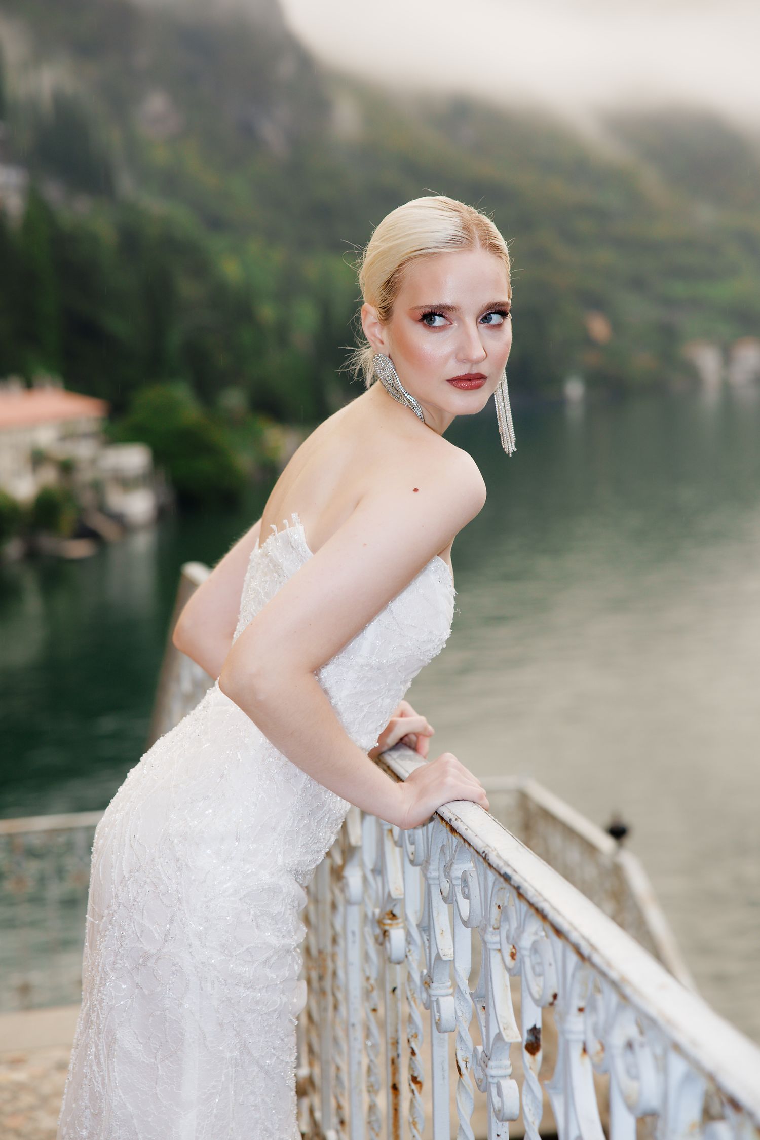 A bride in a white lace gown poses on a balcony overlooking a misty lake.