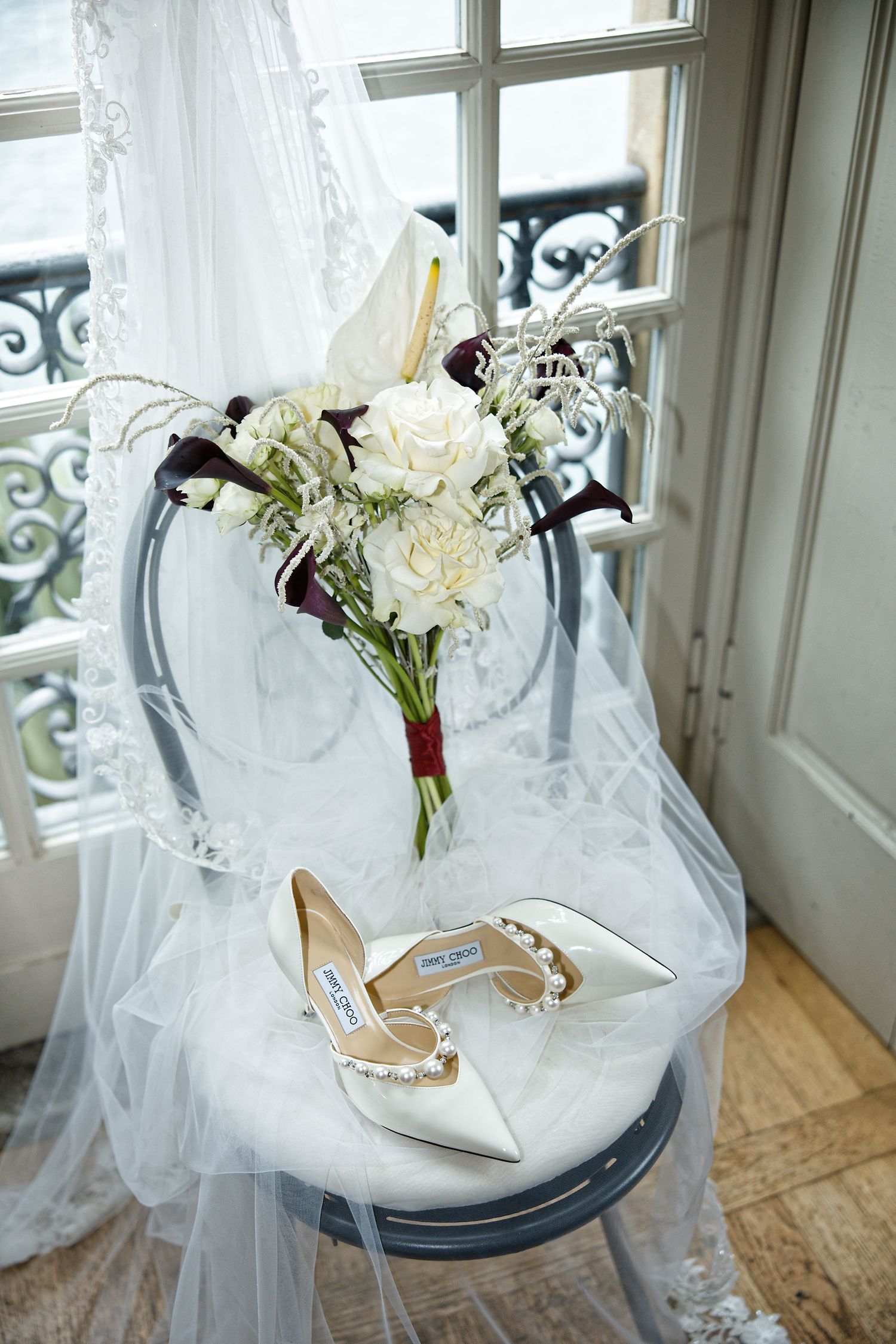 Wedding details featuring a white bouquet and bridal shoes on tulle near a window.