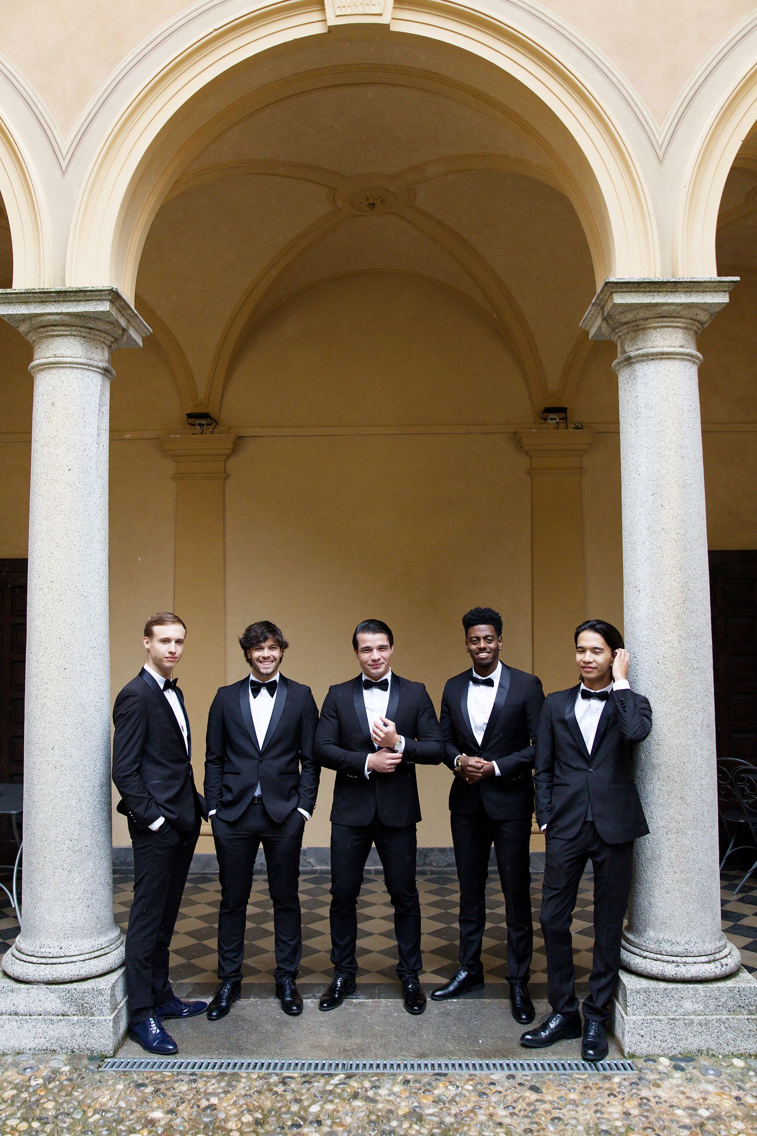 Five groomsmen in black tuxedos and bow ties pose together under elegant arched columns in a formal setting.