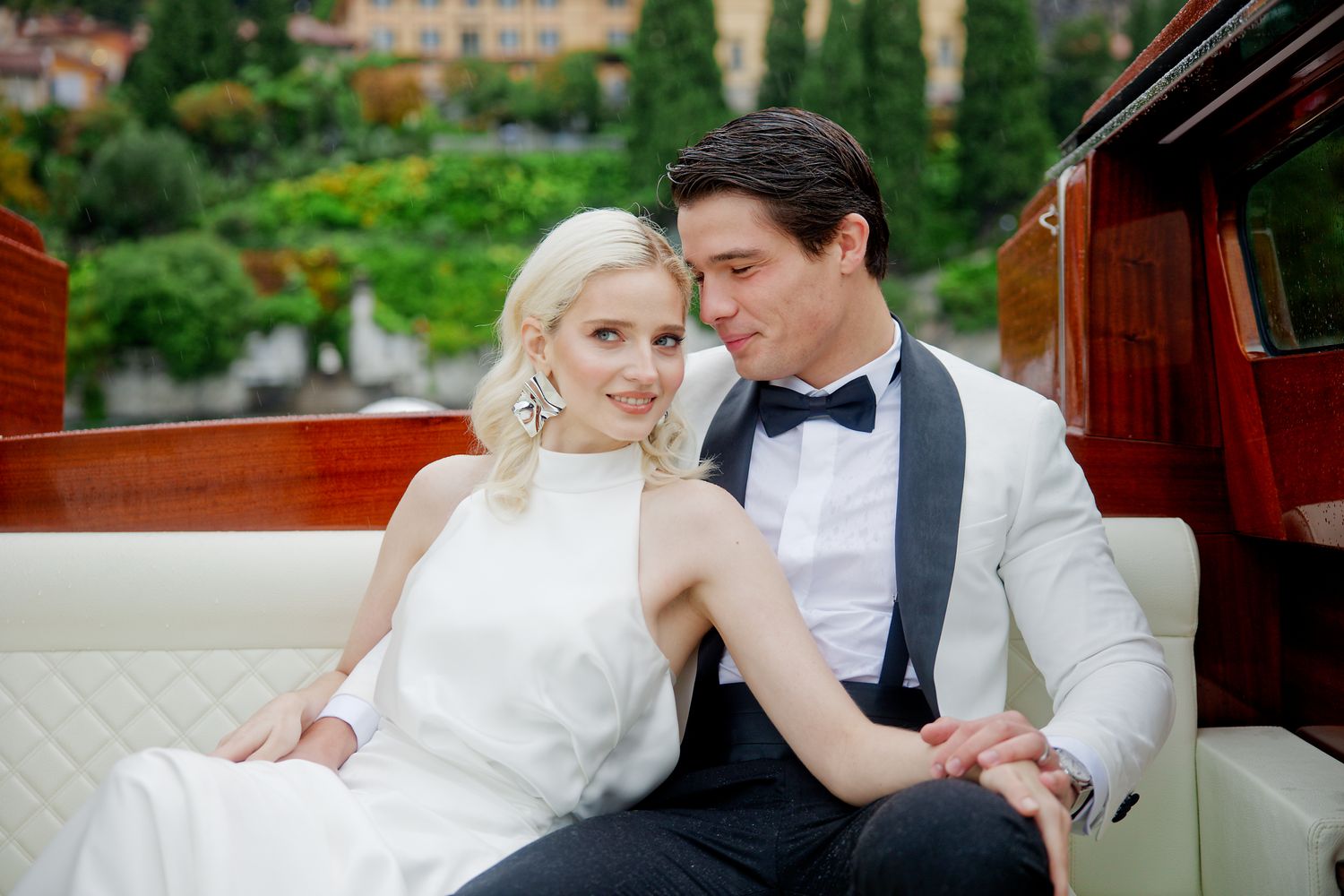 Elegant couple in formal white attire pose together on cushioned boat seating with scenic backdrop.