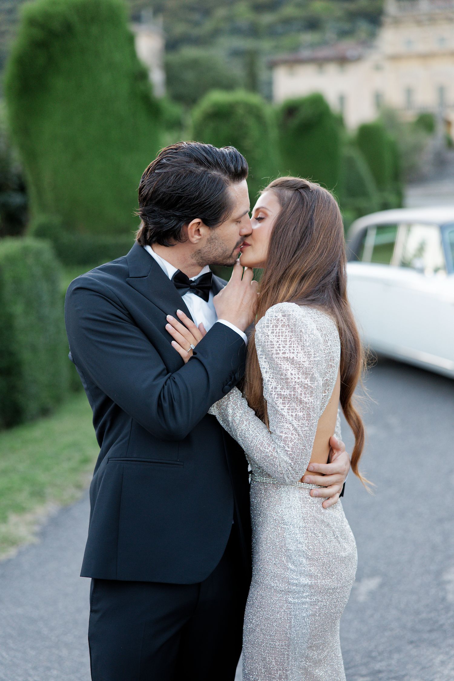 Romantic wedding moment captured on a driveway with classic car and manicured hedges.