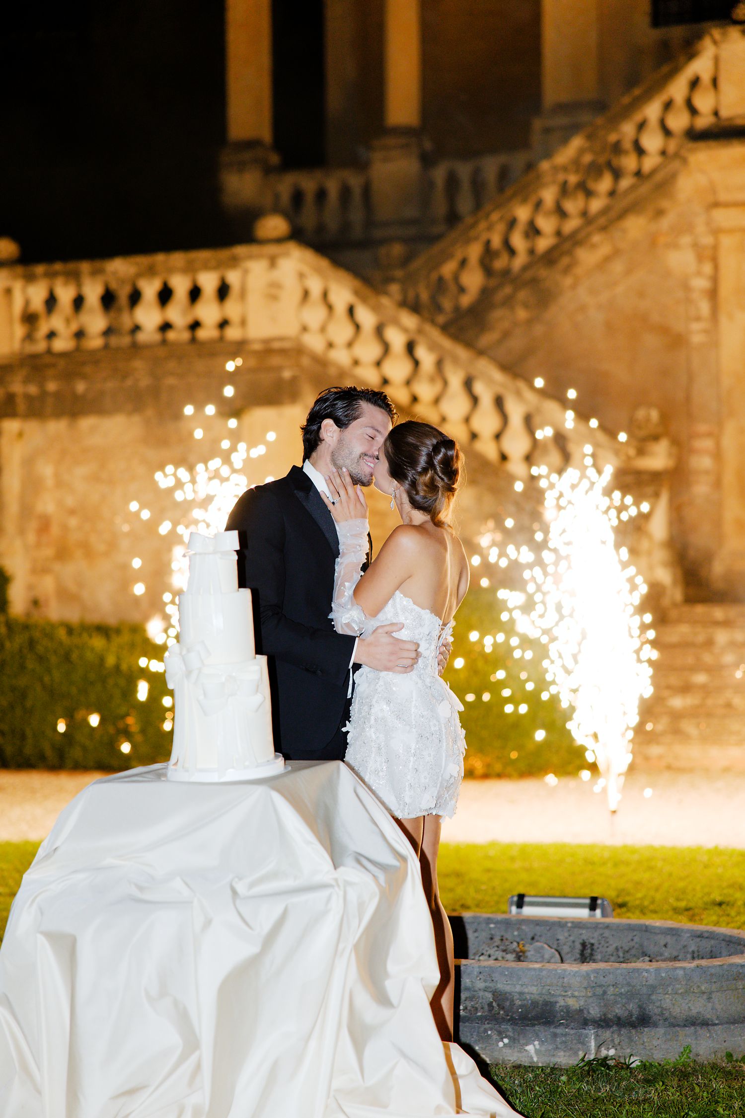 Newlyweds kiss beside their wedding cake as sparklers create a magical backdrop.