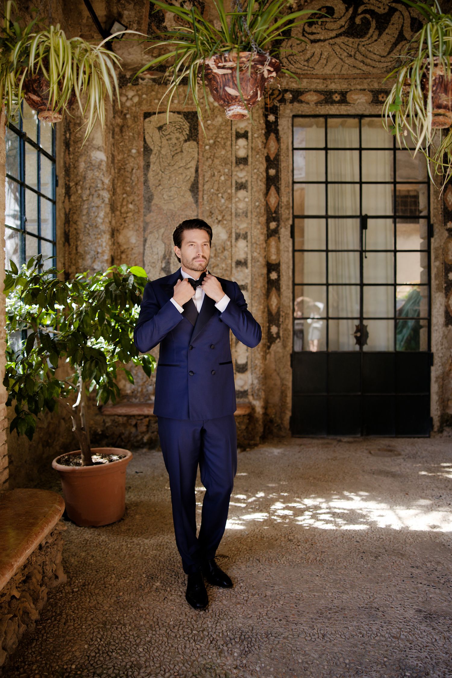 A groom in a navy suit adjusts his bowtie while standing in an elegant venue with hanging plants.