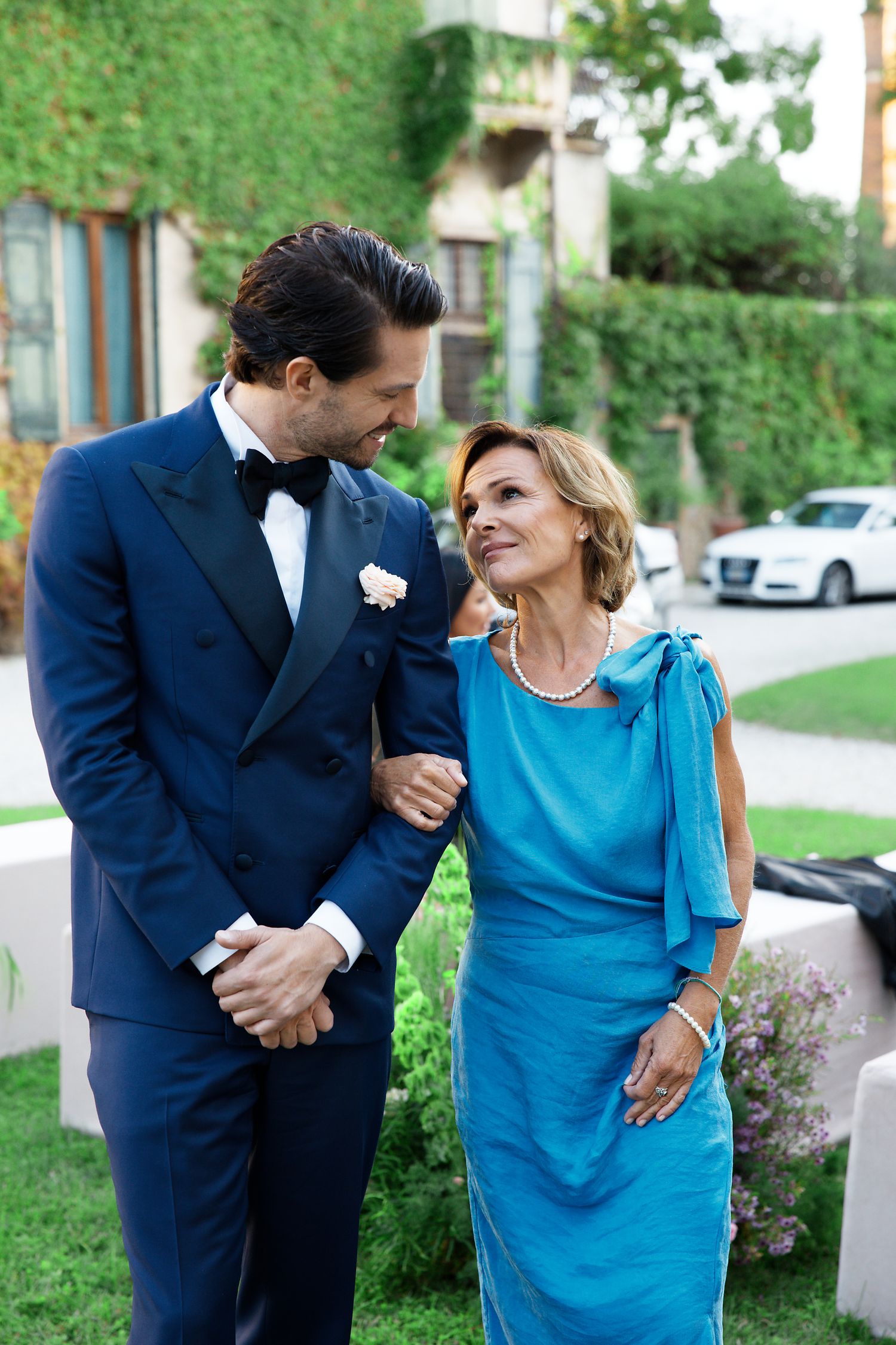 Two people in formal attire share a tender moment at an outdoor garden event, with one wearing navy and one in turquoise blue.