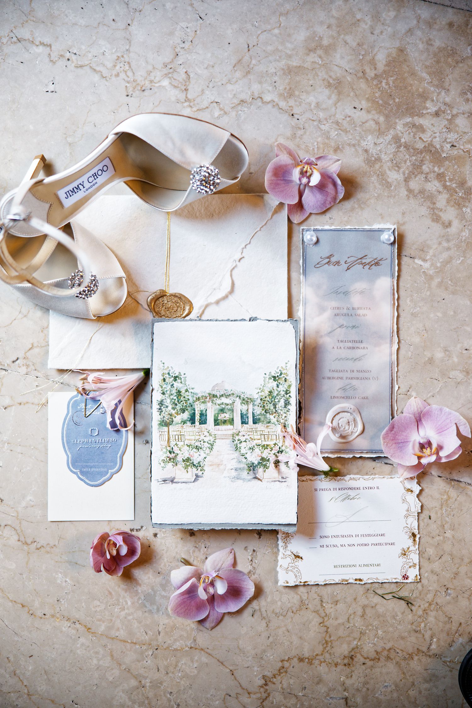 Wedding invitation suite with white bridal shoes, pink orchids, and watercolor venue illustrations on textured surface.