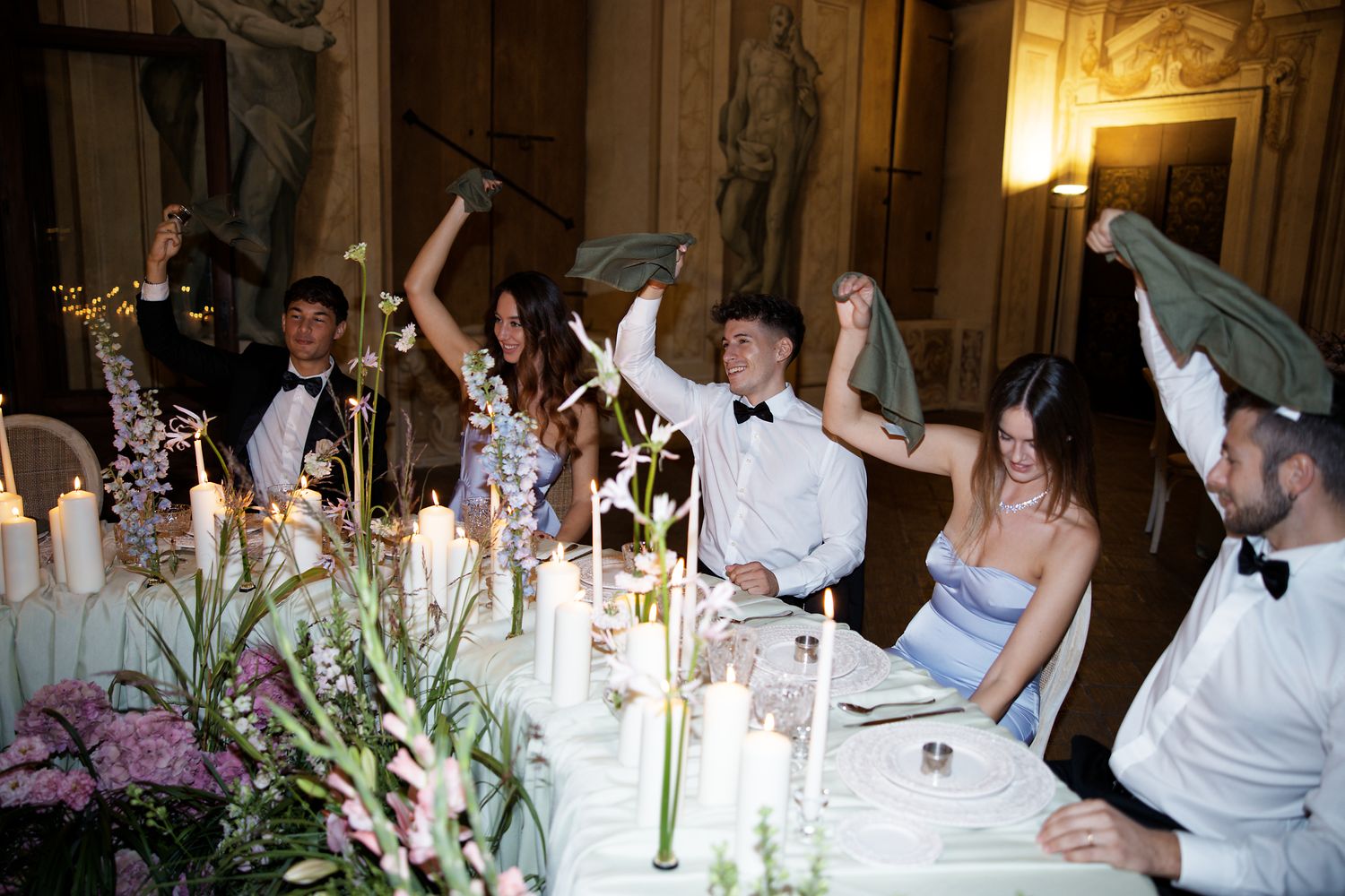 Wedding reception guests raising napkins in celebration at candlelit dinner table.