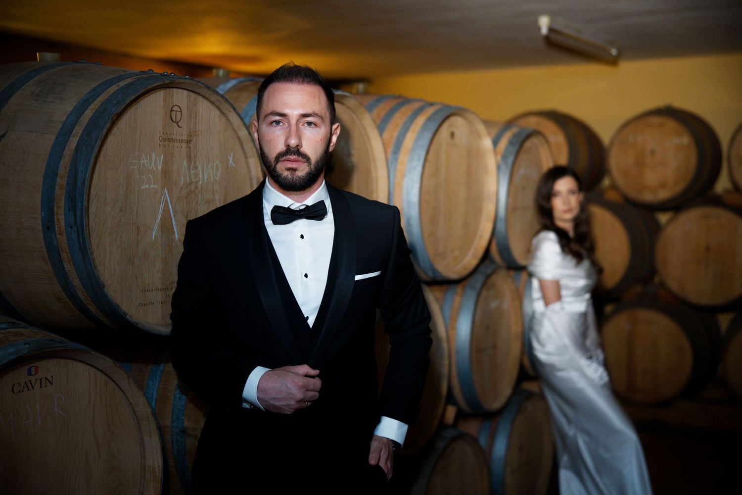 Formal portrait in wine cellar with rustic wooden barrels creating dramatic ambiance.