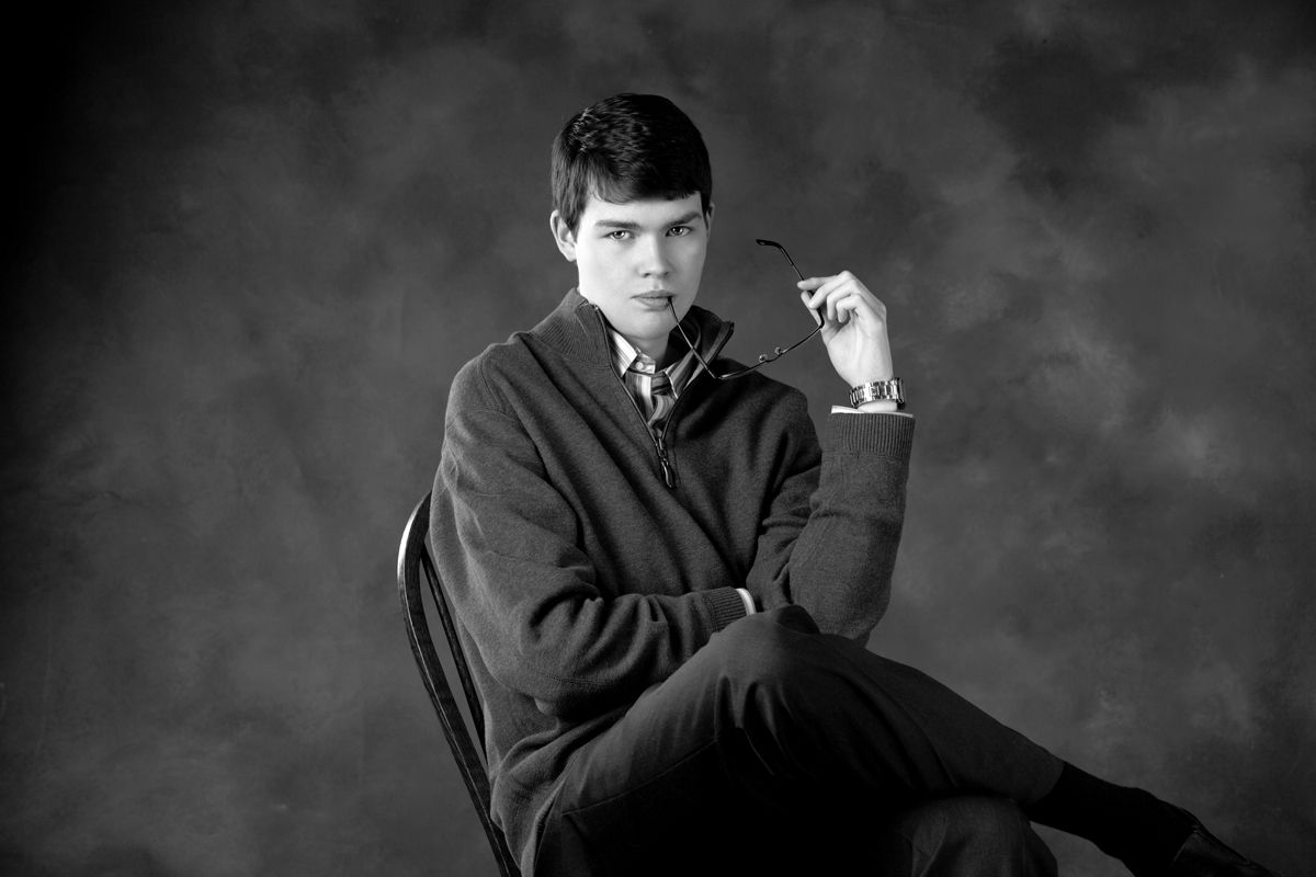 Black and white portrait of a young man seated, holding glasses, wearing a cardigan, against a studio backdrop.