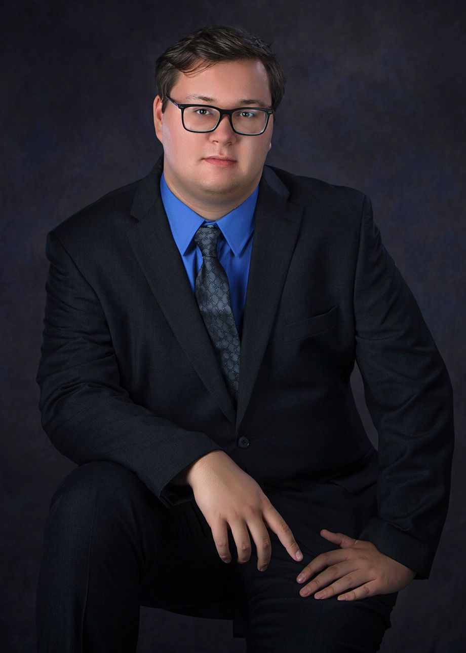 Young man in black suit, blue shirt, and tie with glasses poses for a senior portrait photo, by Jocelyn M. Photography in Rochester, MI
