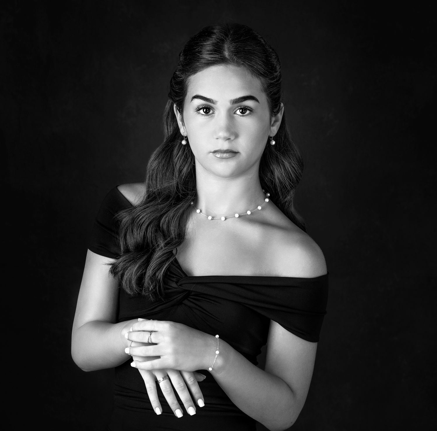 Elegant young woman in black off-shoulder dress with pearl jewelry poses for a classic black and white portrait photo.