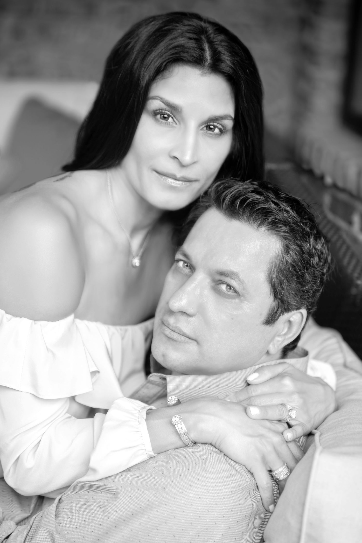 Black and white portrait of an elegant couple posing intimately together, woman in off-shoulder top embracing man from behind.