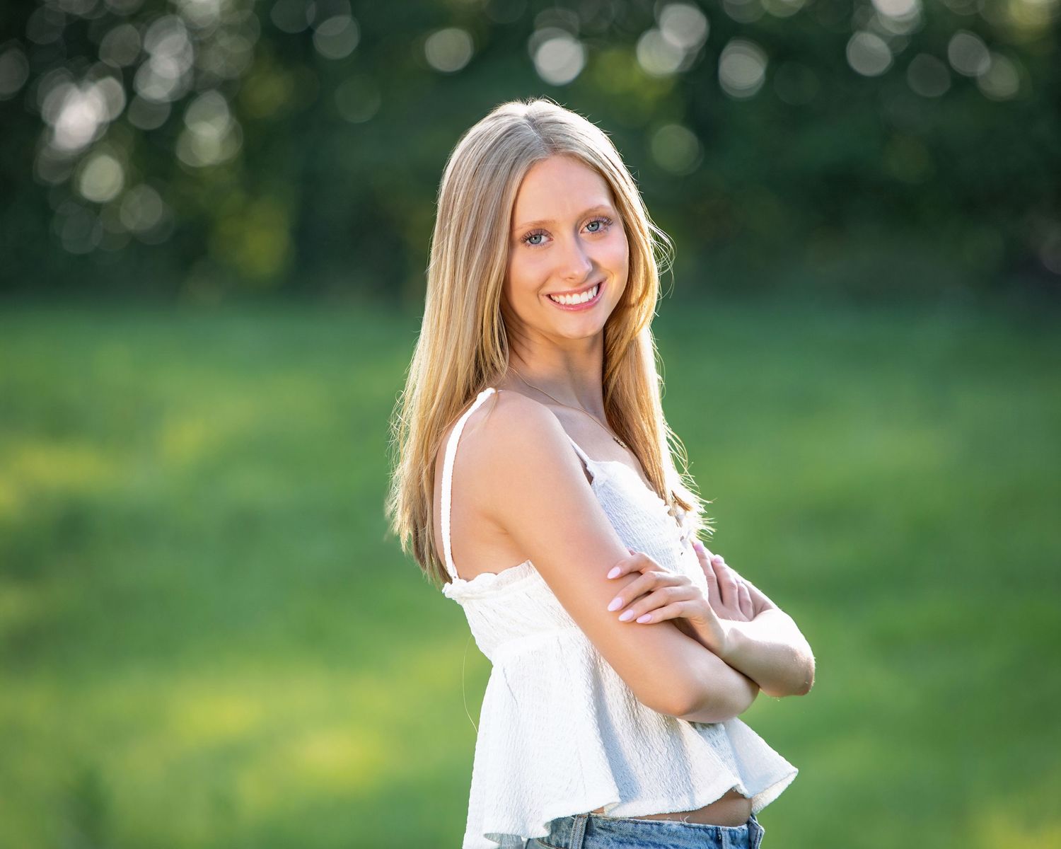 Smiling blonde teenage girl in white top with arms crossed posing outdoors in green field for senior portrait.