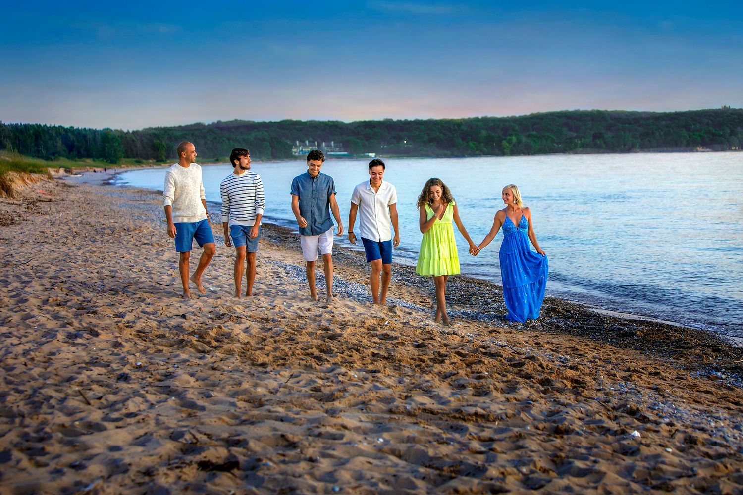 Group of six friends walking along a sandy beach at sunset, wearing summer outfits near calm blue water.