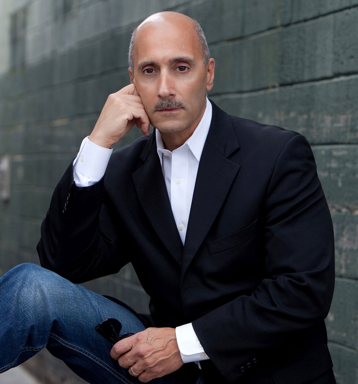 Distinguished bald man with mustache in black blazer seated casually against a green wall.