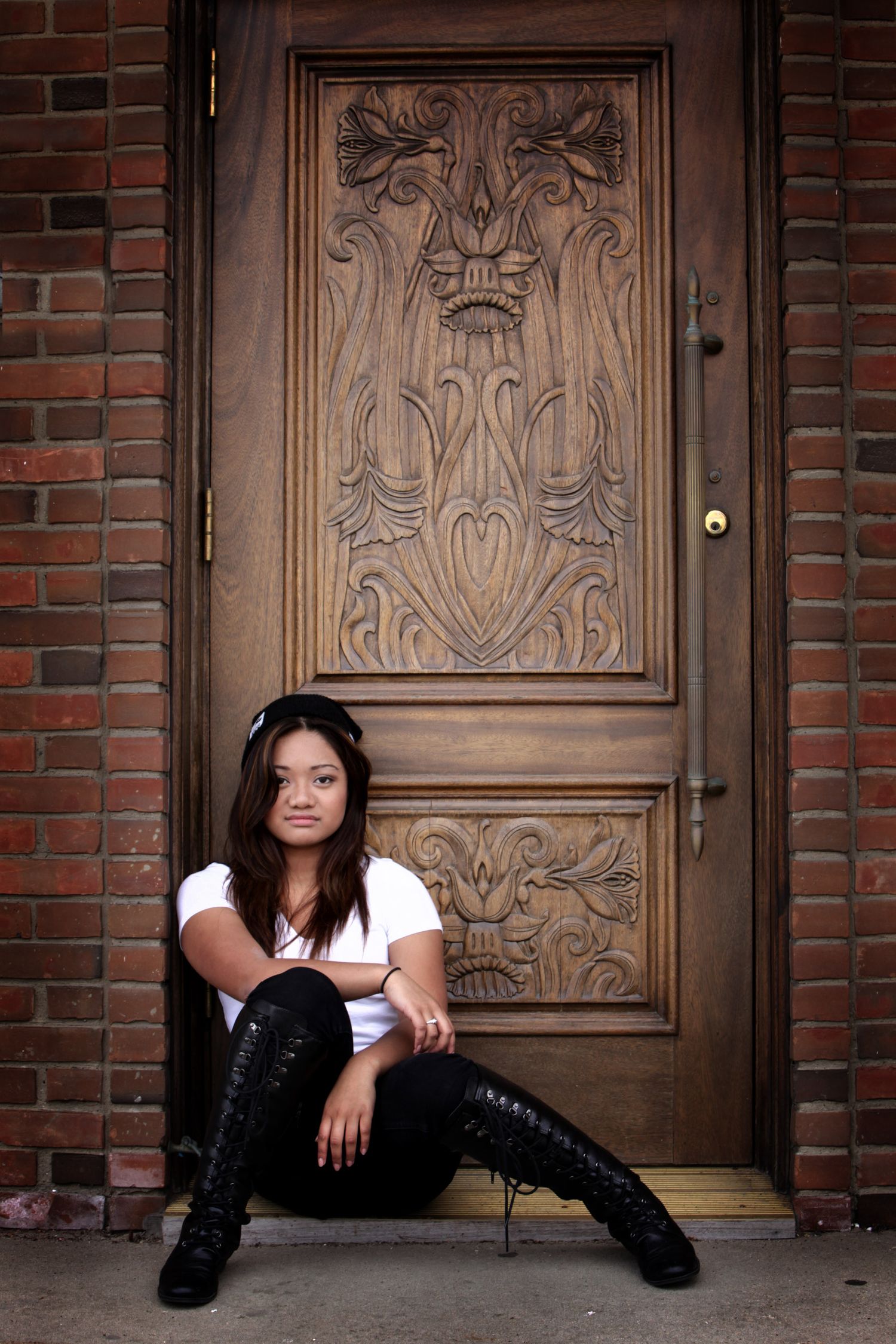 Young woman in black boots and headphones sitting in front of an ornate carved wooden door on brick building.