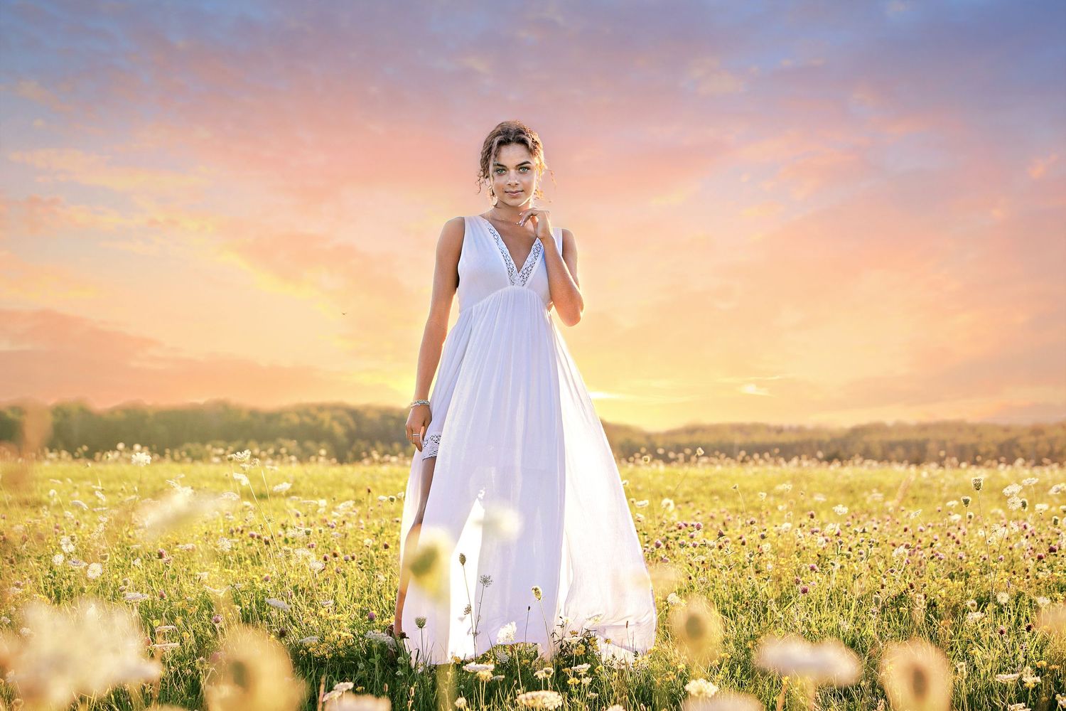 A woman in a flowing white dress stands gracefully in a sunlit field of wildflowers under a pastel sunset sky.
