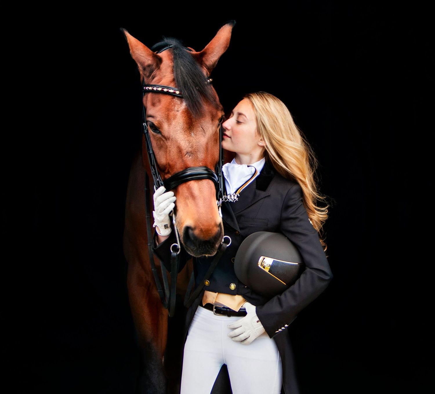 Blonde equestrian rider in black jacket holding helmet while affectionately kissing her bay horse against dark background.