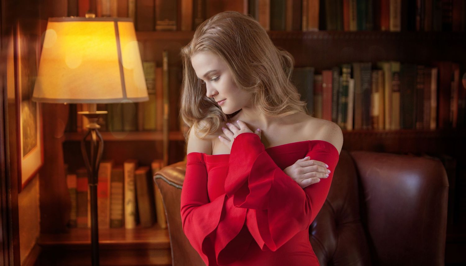 Elegant woman in red off-shoulder dress reading in a cozy library with warm lamp light and bookshelves.