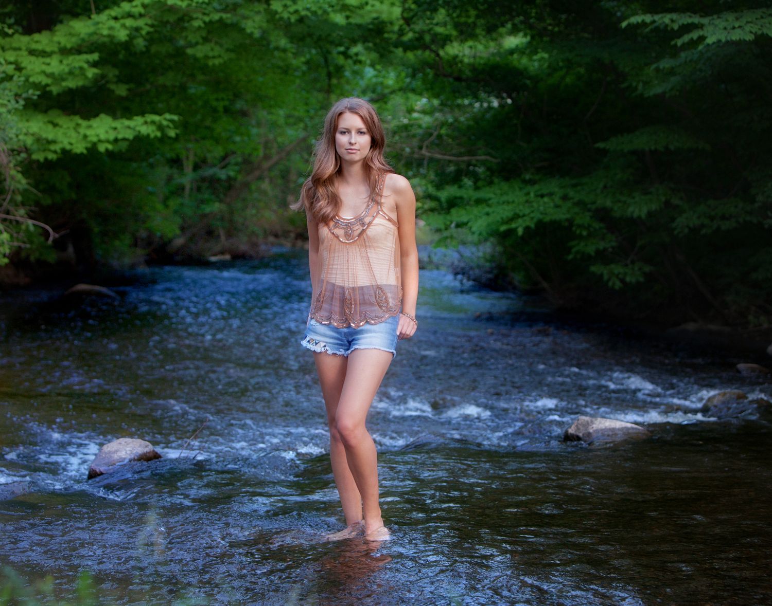 Senior girl in denim shorts wading barefoot through a shallow forest creek in summer,by Jocelyn M. Photography in Rochester, MI
