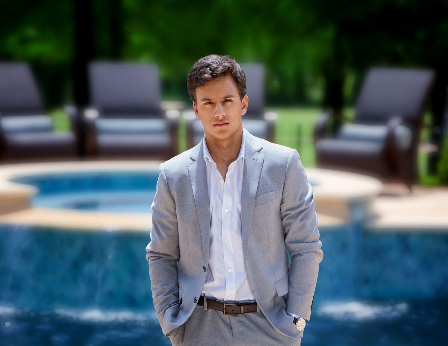 Young man in gray blazer and white shirt stands confidently by a pool with outdoor seating in background.