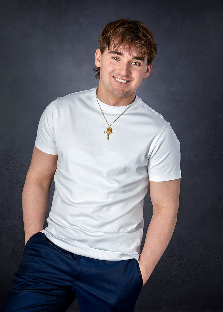 Smiling young man in white t-shirt and gold cross necklace poses for professional portrait.