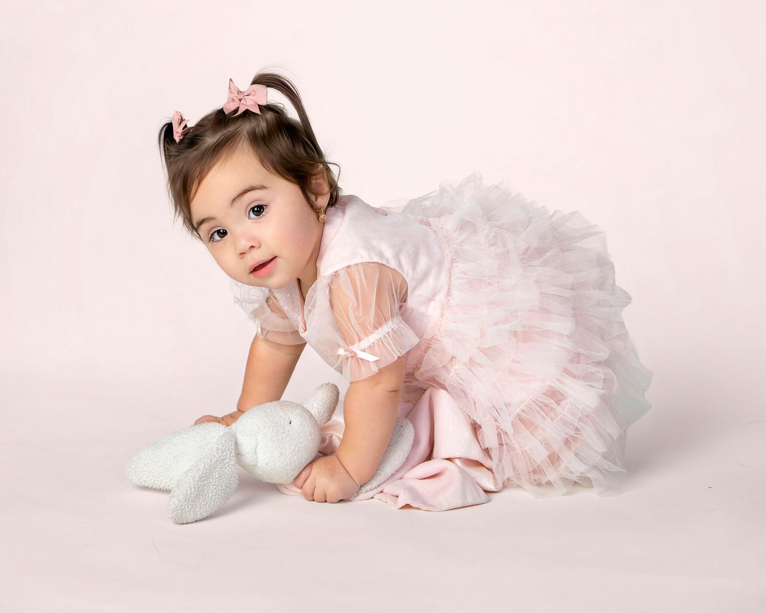 An adorable toddler girl in a white tutu dress and booties poses on a soft pink studio background.