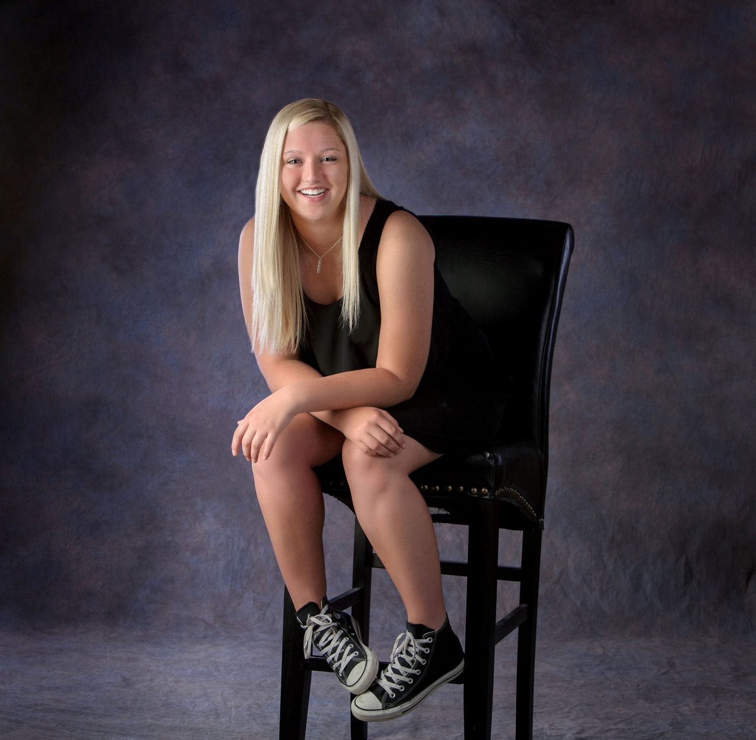 Smiling blonde teen sitting on a black chair in a studio portrait session.