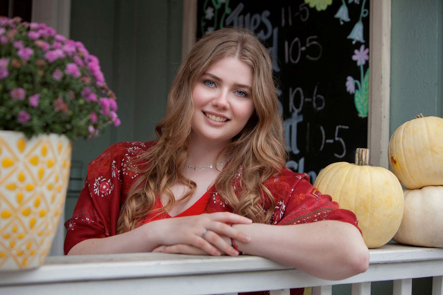 Smiling young woman in red dress leaning on railing with pumpkins and flowers nearby.