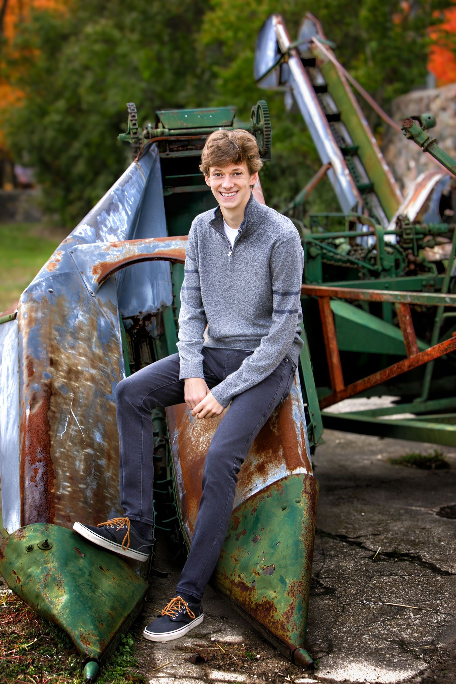 Smiling teen boy in gray sweater sitting on rusty vintage farm equipment outdoors in autumn.