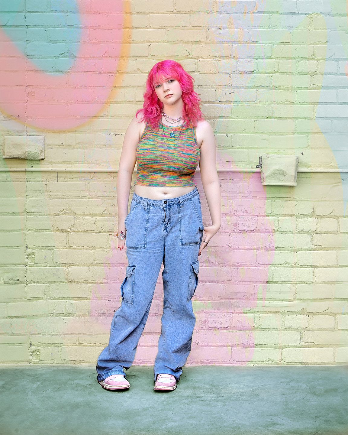 Young woman with pink hair wearing colorful crop top and cargo jeans posing by painted brick wall.