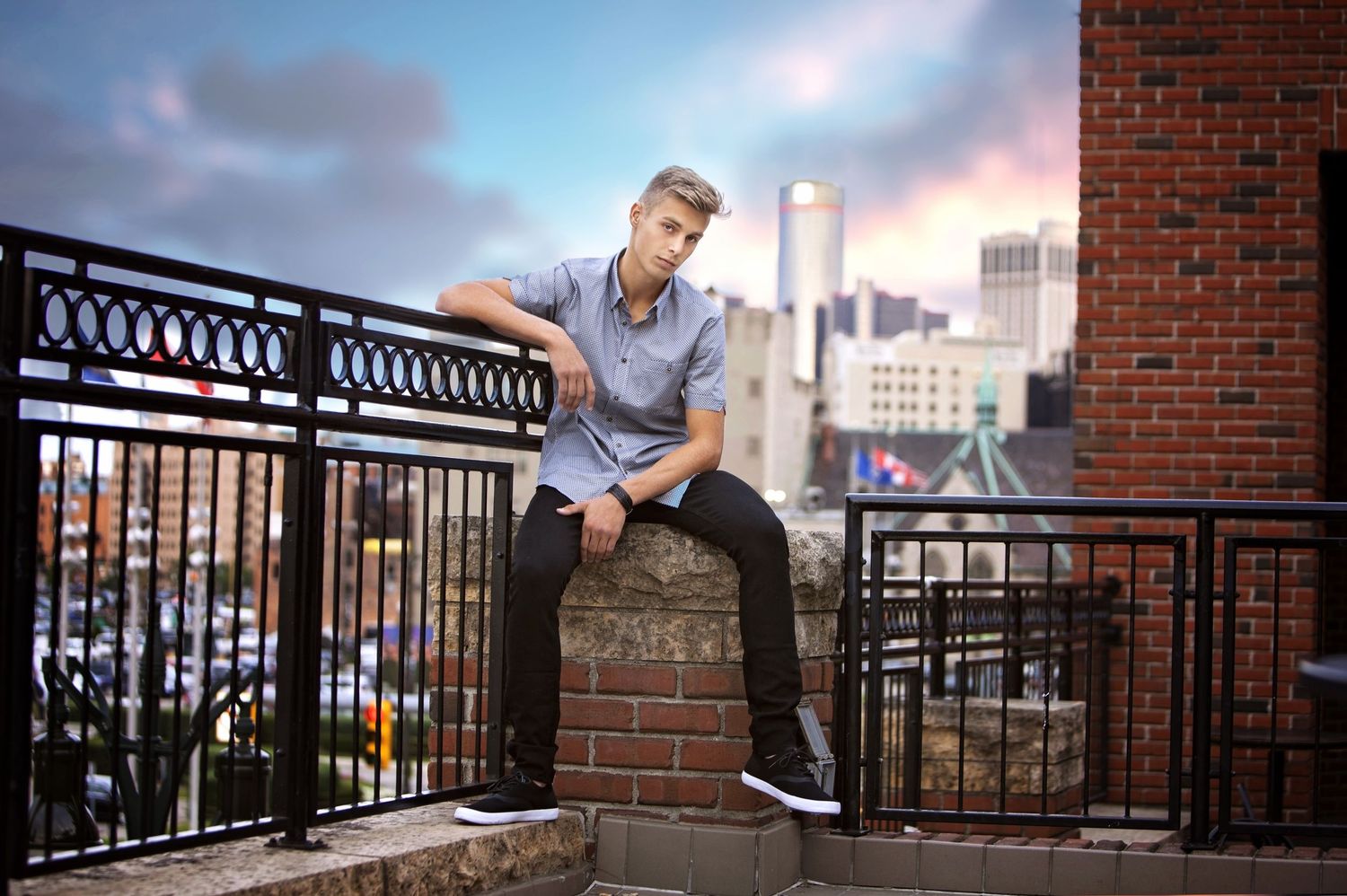 Urban senior session in Rochester, MI by Jocelyn M. Photography—young man sitting on a brick wall with a city skyline.