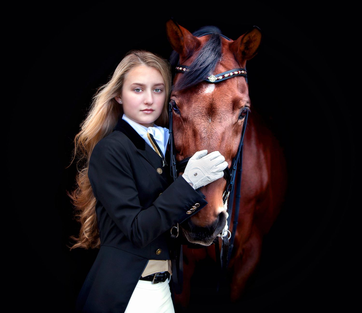 Young equestrian girl in black riding jacket poses with chestnut horse against dark background.
