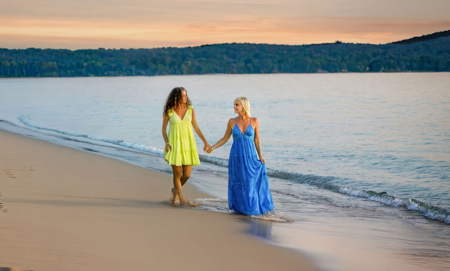 Two women in colorful dresses hold hands walking along a sandy beach at sunset.