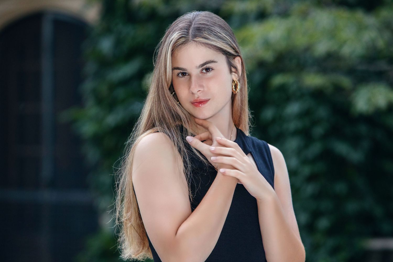 Young woman with long blonde hair in a black sleeveless top posing outdoors with green foliage background.