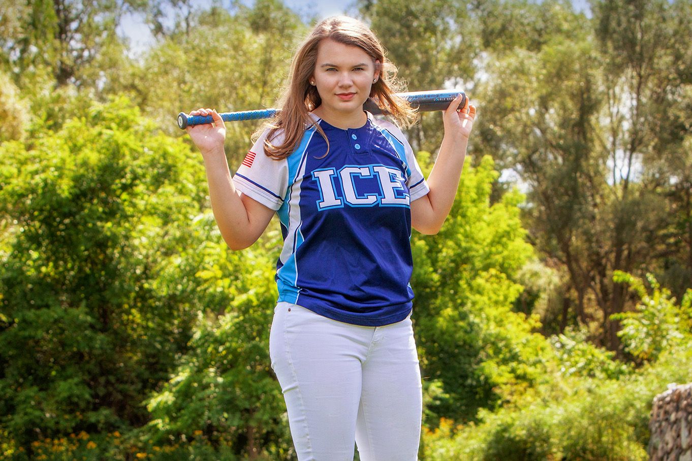 Senior girl portrait baseball jersey holds a bat outdoors in a sunny sports portrait photo, by Jocelyn M. Photography in Rochester, MI