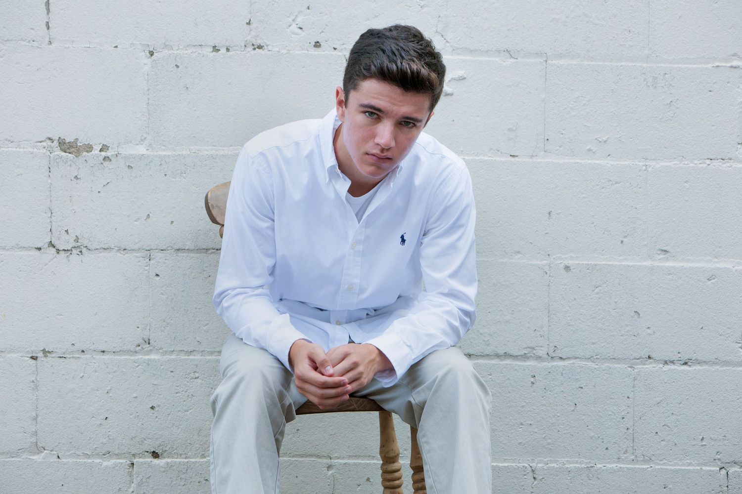 Young man in white button-down shirt sitting on wooden stool against white brick wall, by Jocelyn M. Photography in Rochester,