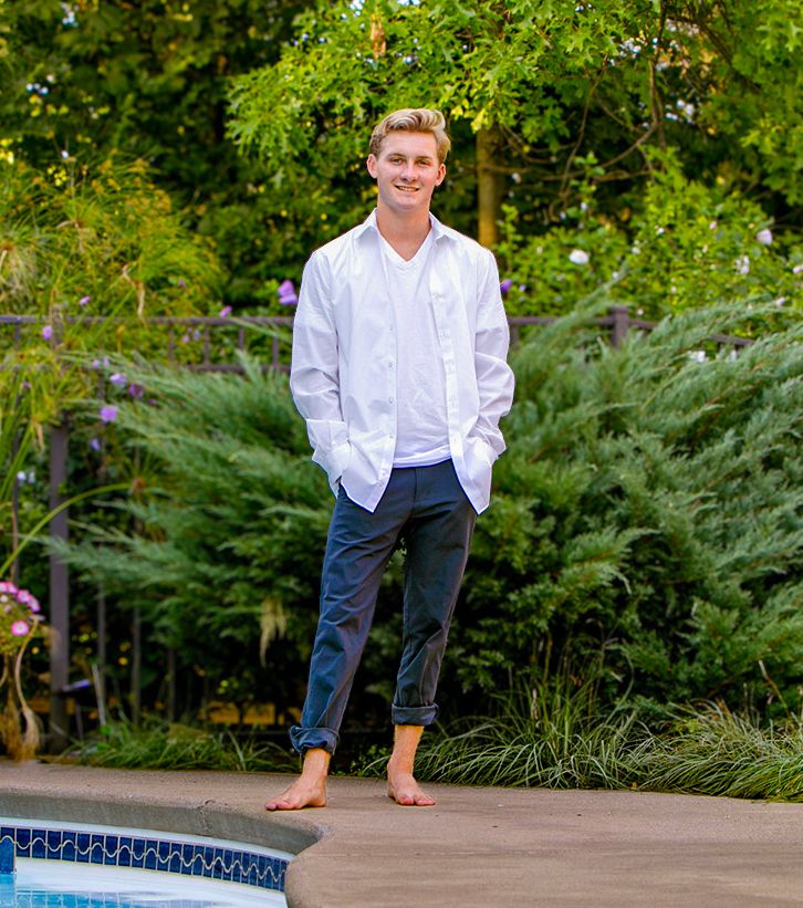 Smiling young man in white shirt and navy pants stands barefoot beside a backyard pool.