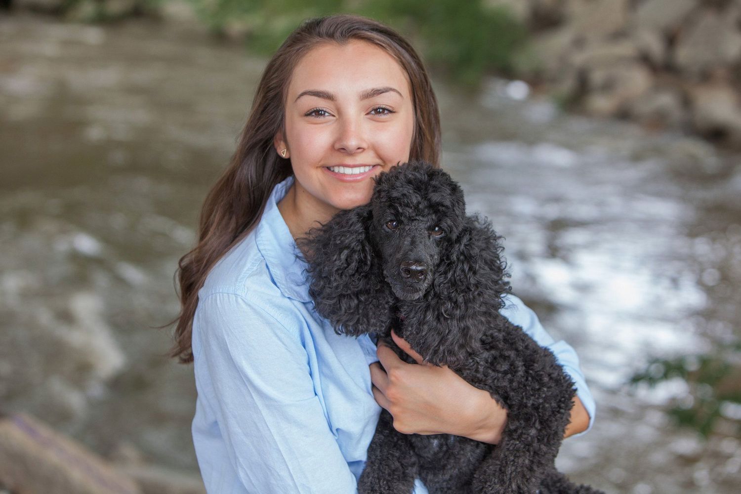 Smiling young woman holding a black poodle near a rocky riverside outdoor setting.