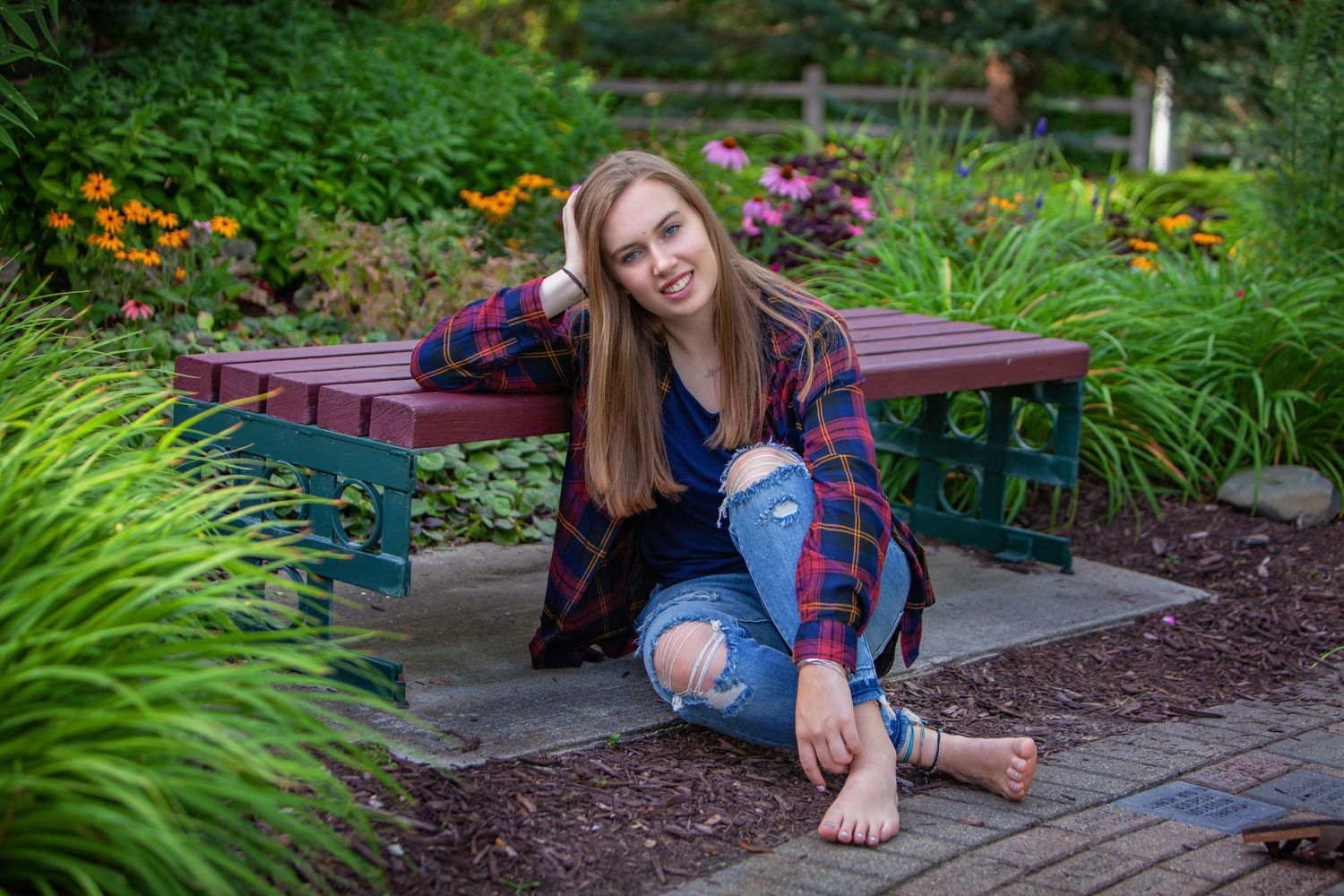 Smiling teenage girl in plaid shirt sitting barefoot beside garden bench surrounded by flowers.