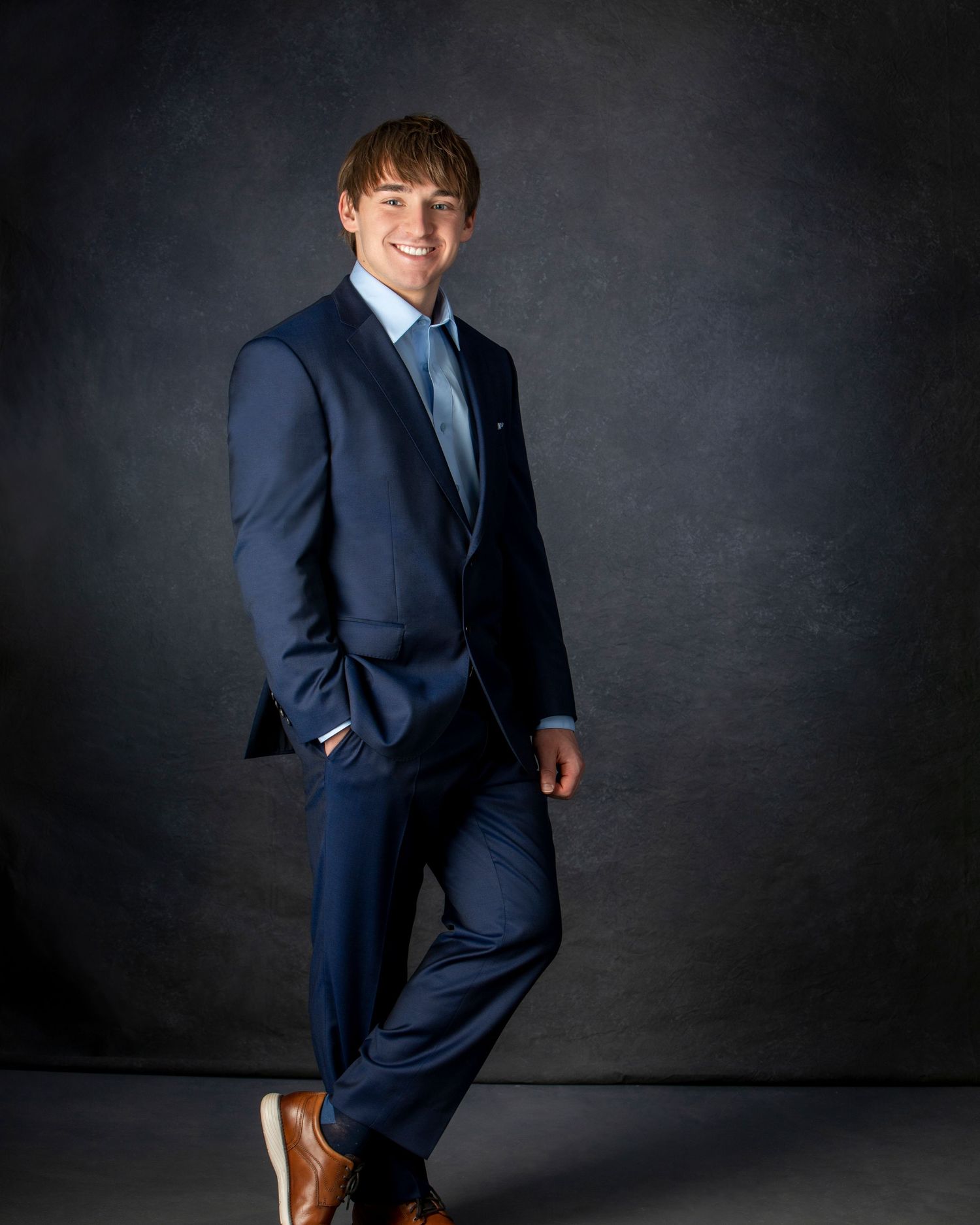 Young man in navy blue suit with light blue shirt and brown shoes poses confidently in professional portrait photo.