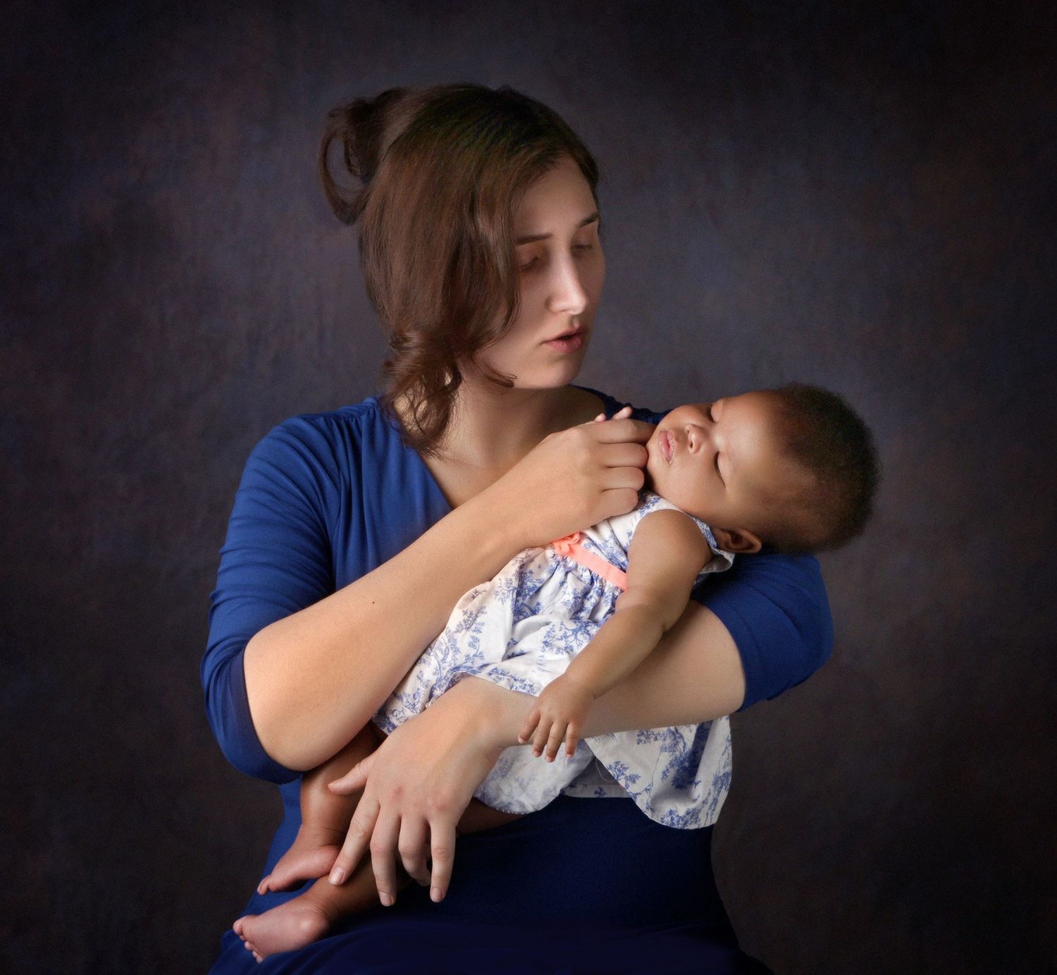 Young mother in blue top tenderly holding sleeping newborn baby in studio portrait.