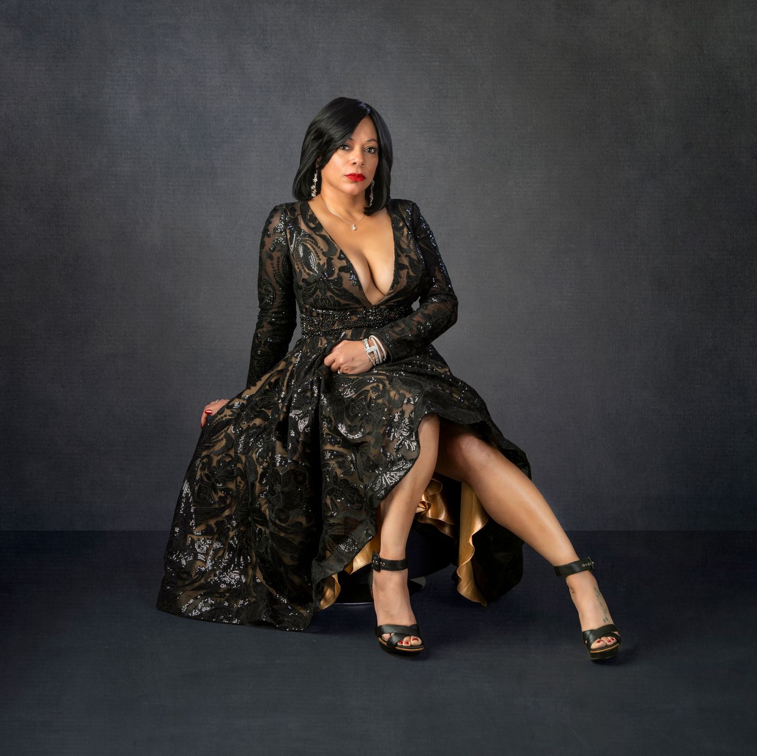 Woman in elegant black lace gown seated on gold stool in professional studio portrait photography.