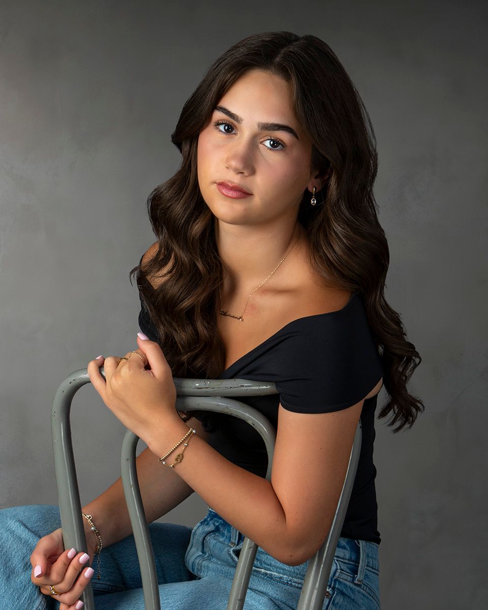 Young woman with wavy dark hair poses gracefully on a metal chair in a black off-shoulder top and jeans.
