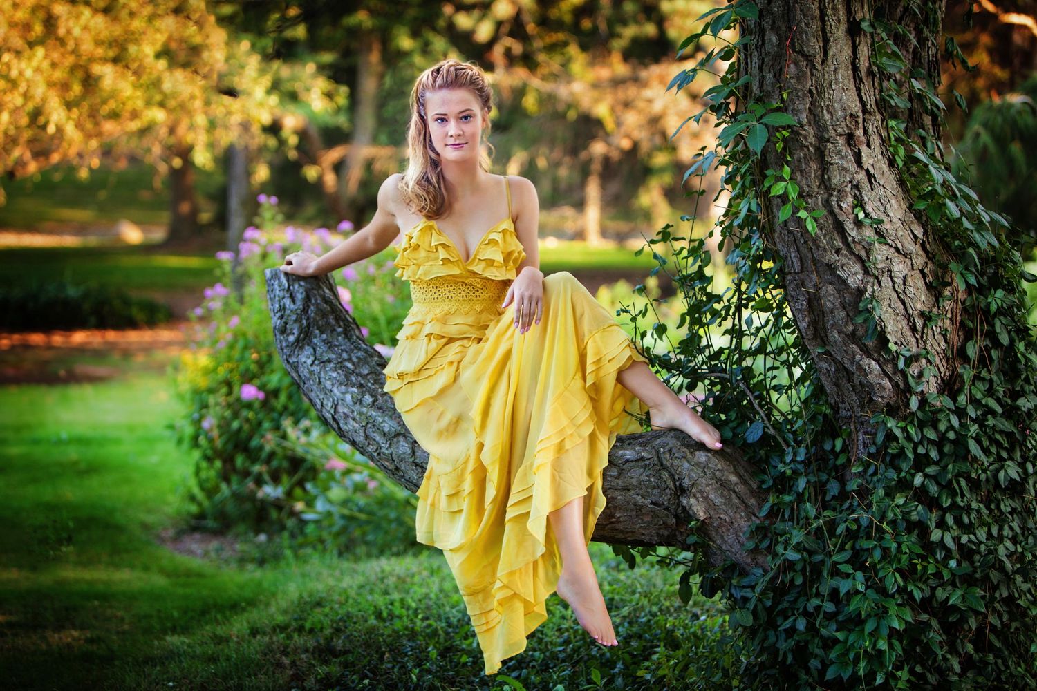 A young woman in a flowing yellow ruffled dress poses gracefully against a tree in a lush green park.
