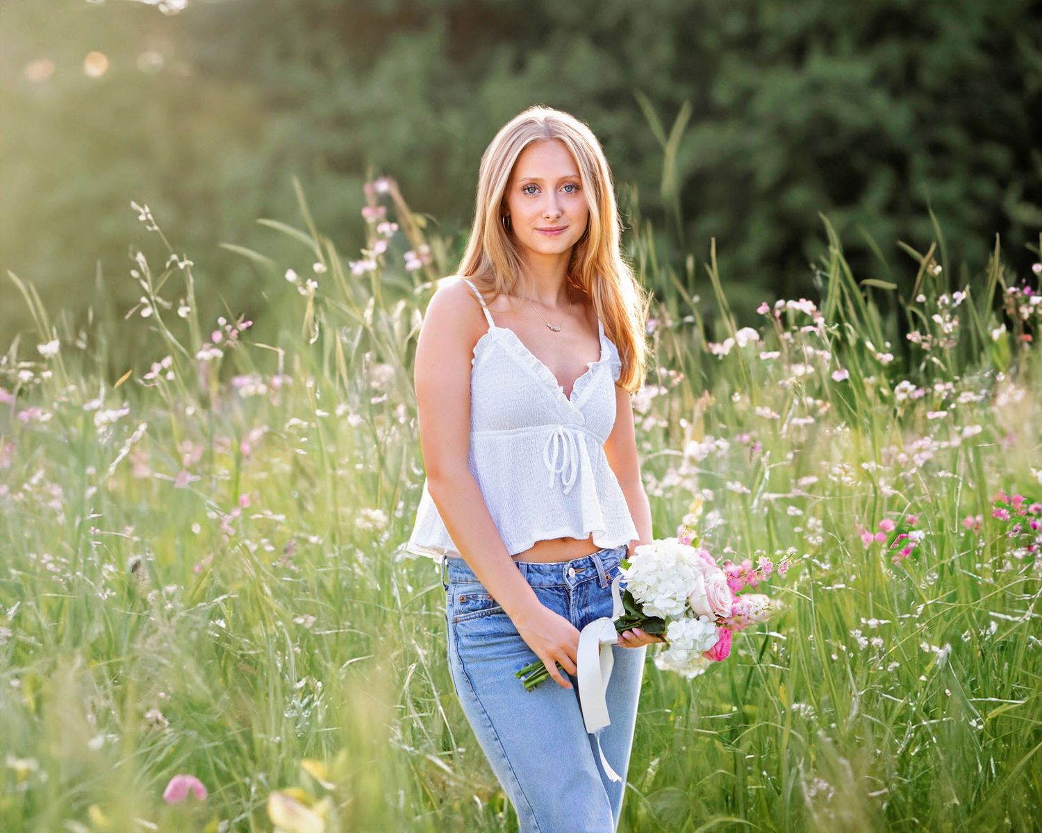 Young blonde woman in white crop top and jeans holding a bouquet of flowers in a sunlit wildflower meadow.